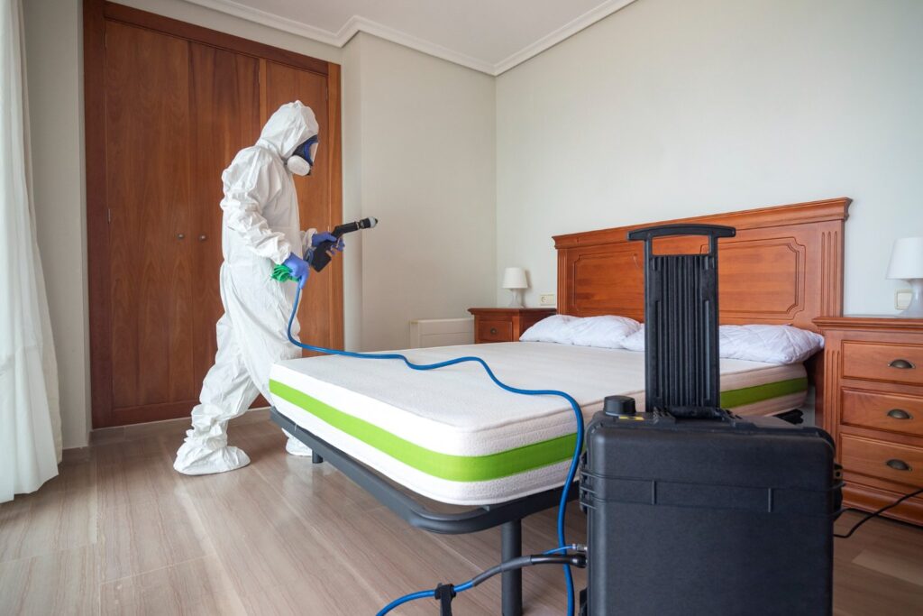 A person in protective gear disinfects a bedroom, spraying a mattress with a cleaning device next to a wooden bed and nightstands.