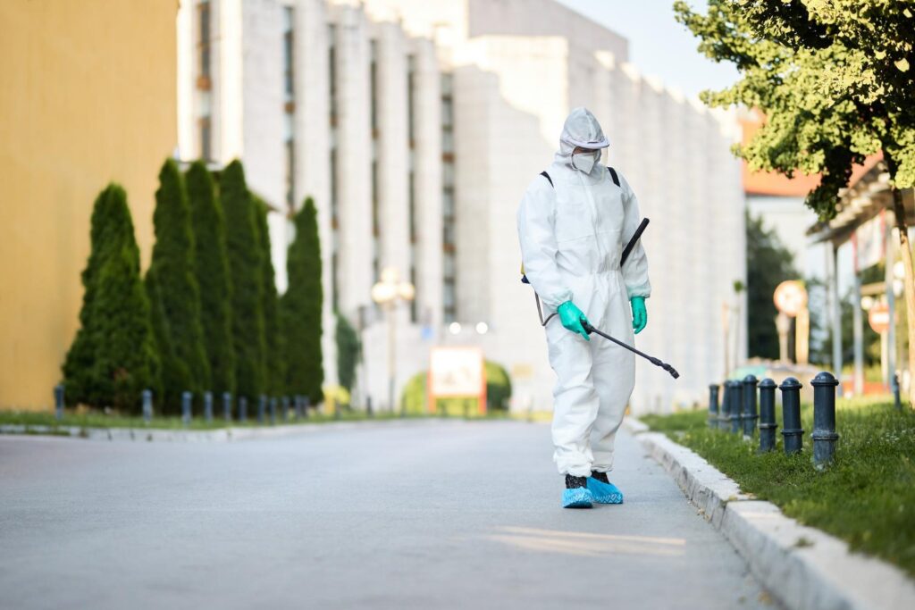 A person in full protective gear sprays disinfectant on a sidewalk along a street, with buildings and trees in the background.